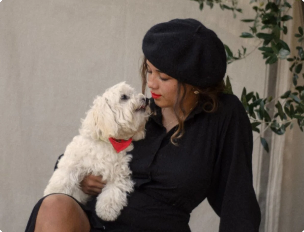 Person in a black beret holds a small white dog wearing a red bandana; they look at each other